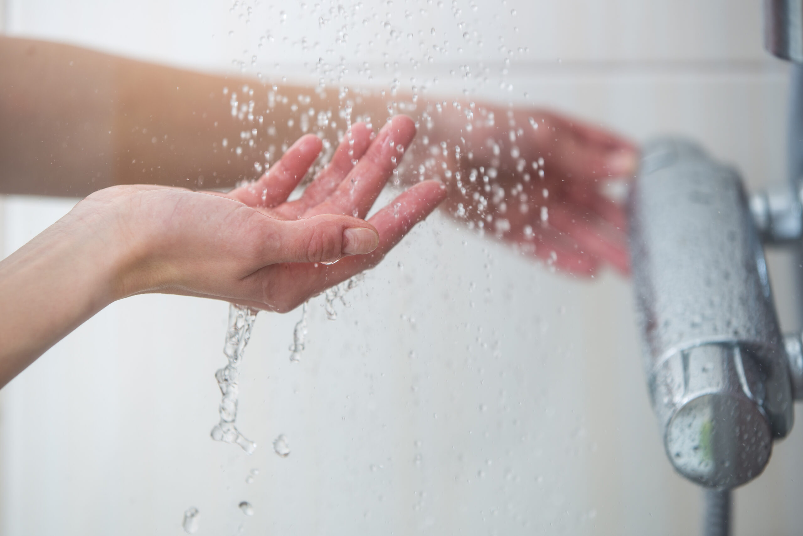 Hands are cupped under running water from a shower faucet, with water droplets visible against a blurred background.