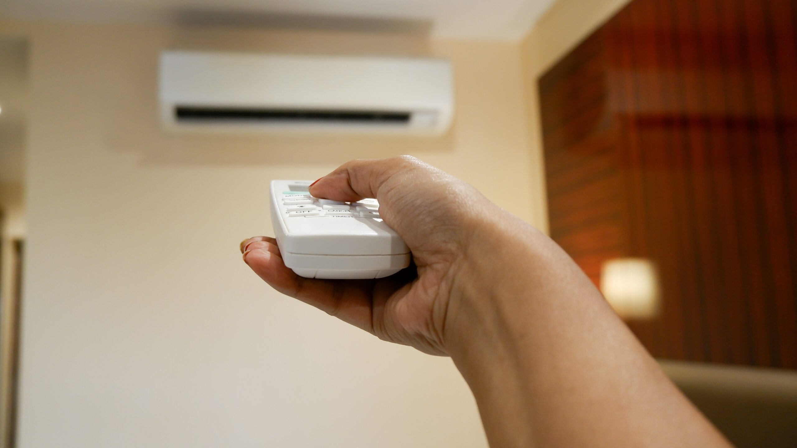 A person points a remote control at a wall-mounted air conditioning unit, contemplating if it's time to schedule both an AC check-up and the overdue water heater repair.