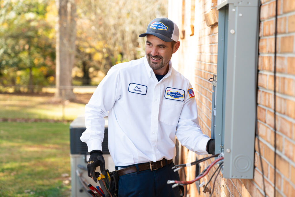 Electrician in uniform works on outdoor electrical panel attached to a brick wall, holding tools, with a grassy area and trees in the background.