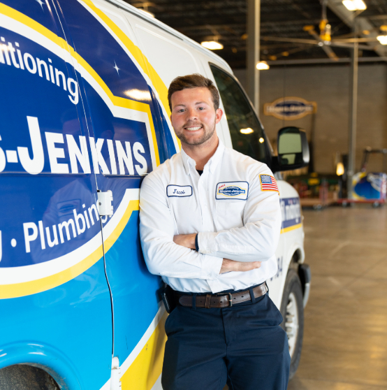 A technician in uniform smiles while leaning against a company van in a warehouse. The van displays a blue and yellow logo.