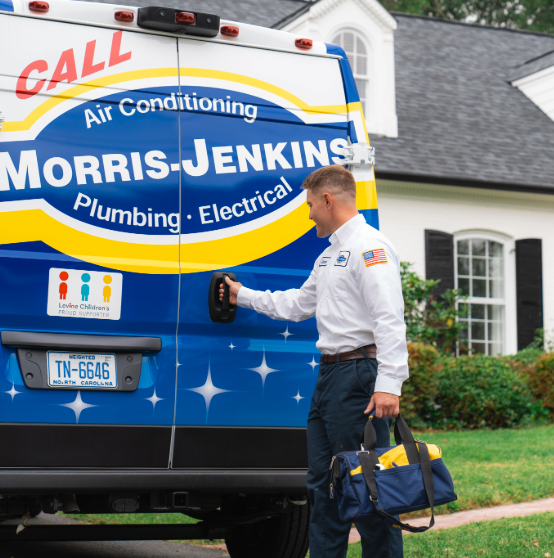A technician stands beside a Morris-Jenkins service van, holding a tool bag outside a house. The van advertises plumbing, electrical, and air conditioning services.