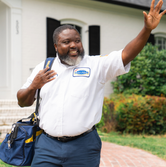 A person in a uniform waves while carrying a bag. A house with steps and greenery is in the background.