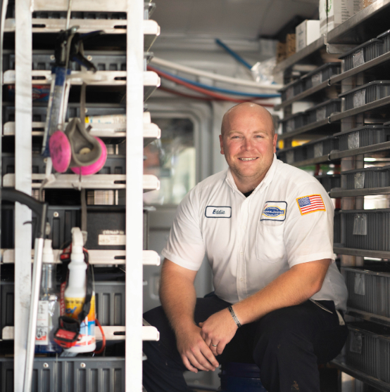 Man in uniform with name tag sits in a vehicle aisle lined with shelves, surrounded by organized tools and supplies.