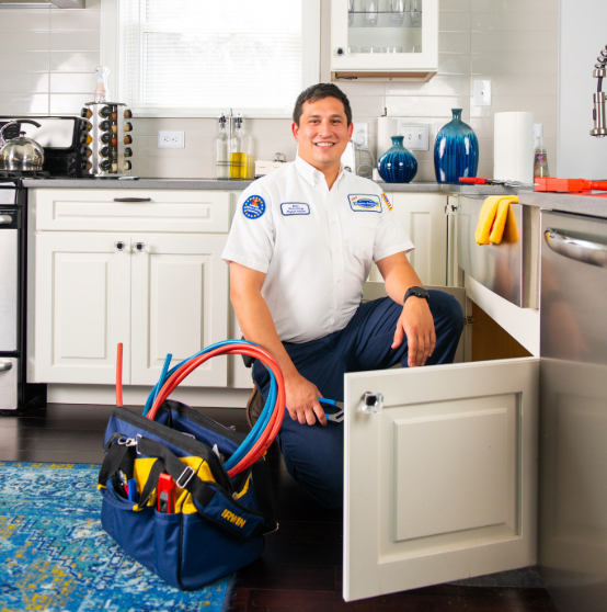 Emergency plumber kneels by an open cabinet in a kitchen, holding a wrench. A tool bag with hoses and tools is on the floor.
