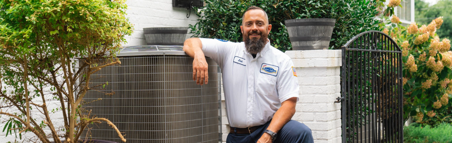 A technician in a uniform kneels beside an outdoor air conditioning unit near a white brick wall and potted plants.