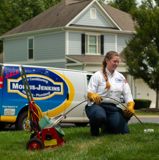 Technician in uniform kneels on grass using equipment, with a service van and a house in the background.