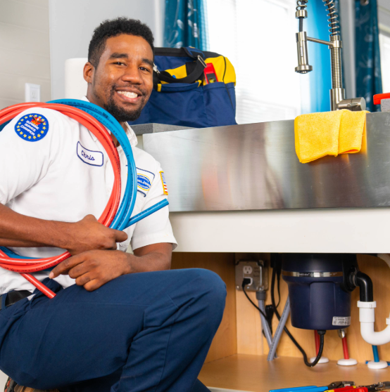 A plumber kneels by a sink, holding red and blue pipes. A tool bag and a yellow cloth are on the counter. He is smiling and wearing a uniform with a name tag.