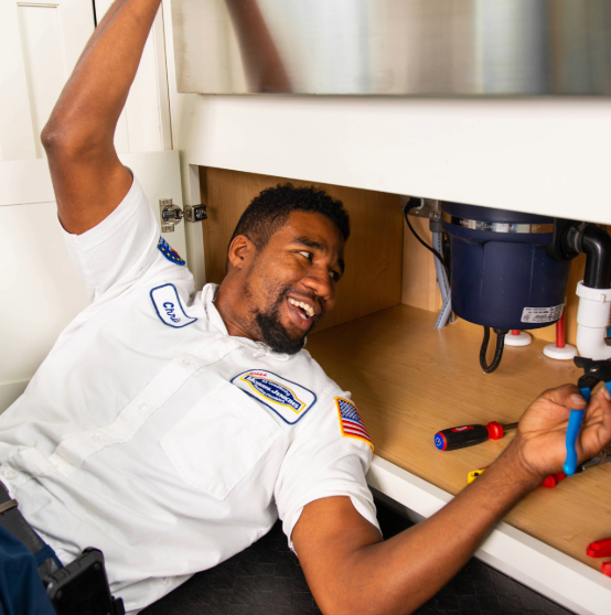 A smiling plumber in a white uniform works under a sink, using pliers, surrounded by tools.