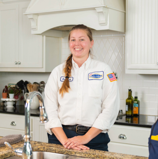 Person in a white uniform with name tag stands in a kitchen by a countertop, smiling. Cabinets and kitchen items are visible in the background.