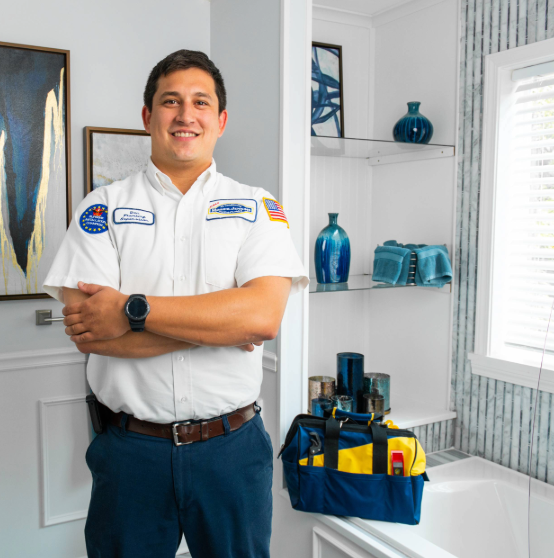 A man with folded arms stands in a bathroom with a blue toolbox on the counter. He wears a white uniform shirt with patches. The bathroom has blue accents and a window with blinds.