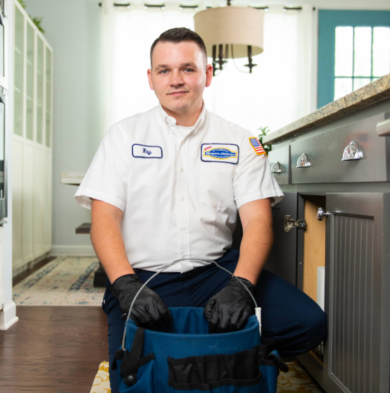 A man in a uniform with black gloves kneels by a sink holding a tool, suggesting he's performing maintenance or repair work.