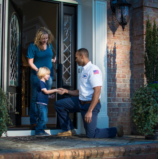 A service technician kneels at the doorway while greeting a young boy, as a woman stands behind them, in front of a brick house.