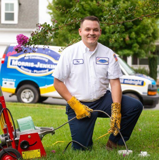 A technician kneels on grass wearing gloves and a uniform, handling a cable machine. A Morris-Jenkins service van is parked in the background.