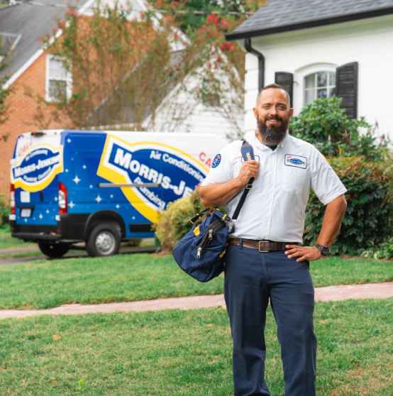 A bearded technician stands on a lawn holding a tool bag in front of a branded service van and a house.