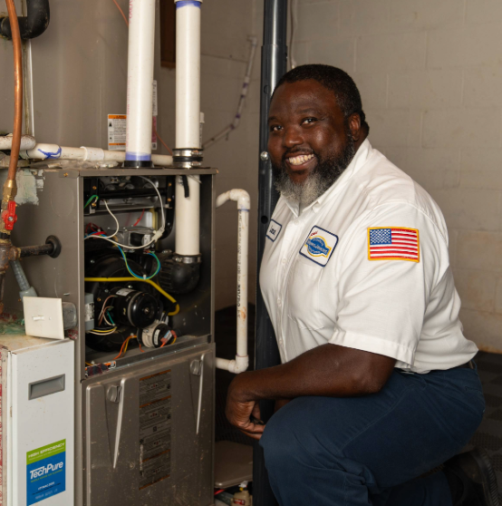 A man in a uniform kneels beside an open HVAC unit in a basement, smiling for the camera.