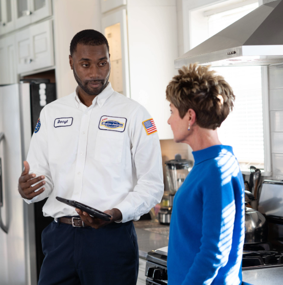 A technician in uniform discusses with a woman in a blue sweater in a kitchen. He holds a tablet, gesturing as they talk.