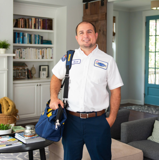 Man in a utility uniform with a name tag stands in a living room, holding a blue bag. The room features bookshelves, a sofa, and a coffee table.