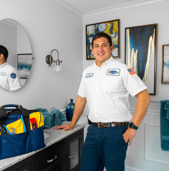 A plumber in a white uniform stands in a bathroom beside a tool bag on a counter. He is smiling and positioned near wall-mounted artwork and lighting.