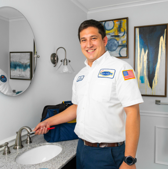 A plumber smiles while holding a wrench over a bathroom sink. He wears a uniform with an American flag patch and stands in a modern bathroom with abstract art on the walls.