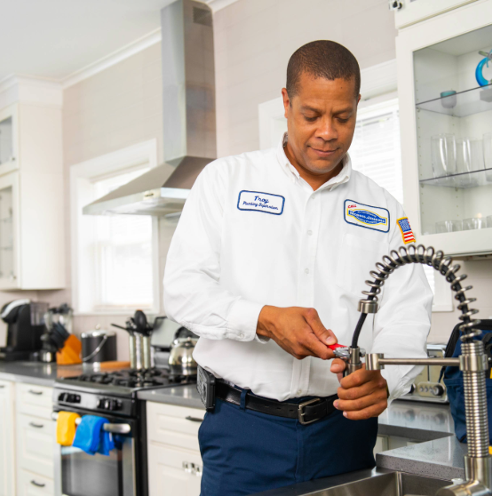 A plumber fixes a kitchen faucet in a modern kitchen, wearing a white uniform with name tag.