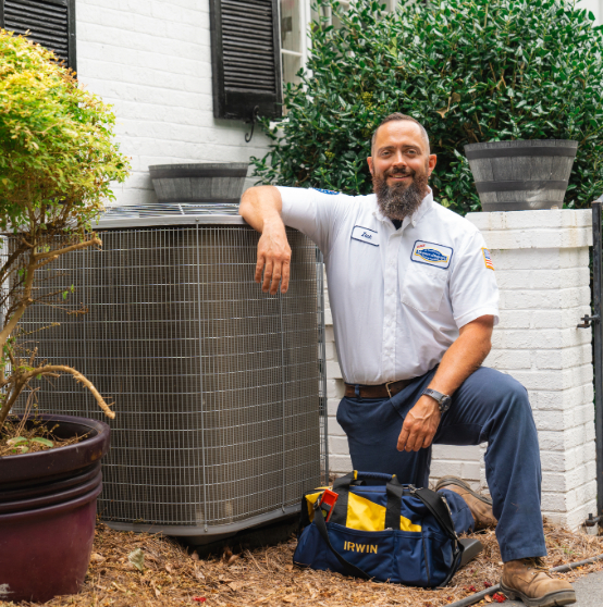 Person in uniform kneeling beside an outdoor air conditioning unit. They are in front of a white brick wall and have a tool bag labeled "IRWIN" at their side.