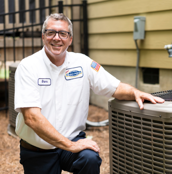 A person in a white shirt with a name tag kneels next to an outdoor air conditioning unit.