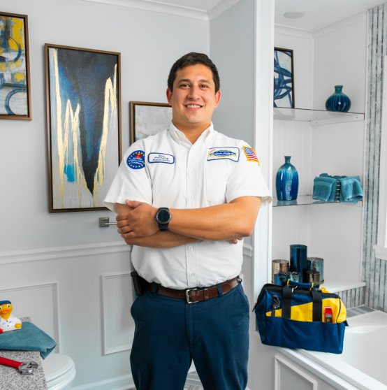 A person in a white uniform stands with arms crossed in a modern bathroom. A tool bag is on a shelf nearby.
