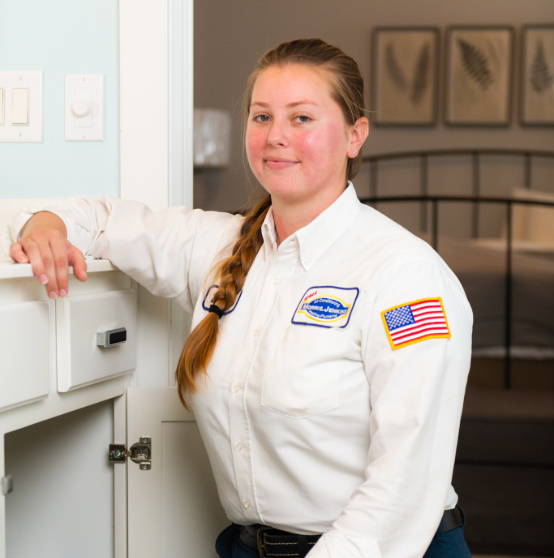 Person in a white uniform with badges, including an American flag, kneeling by a white cabinet in a room with a bed and framed artwork.