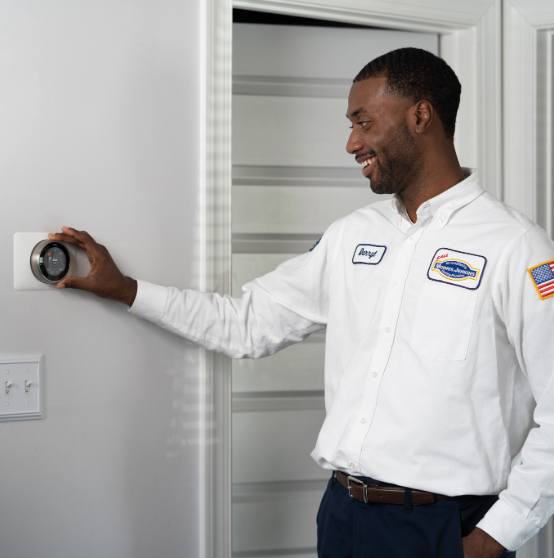 A person in a white uniform adjusts a thermostat on a wall in a home setting.