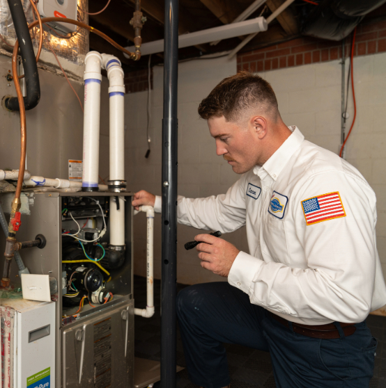 Technician in a uniform inspecting a furnace system, holding a flashlight, in a basement setting.