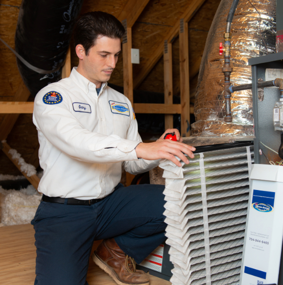 A technician in a white uniform is examining an HVAC filter in an attic space. A large duct and various equipment surround him.