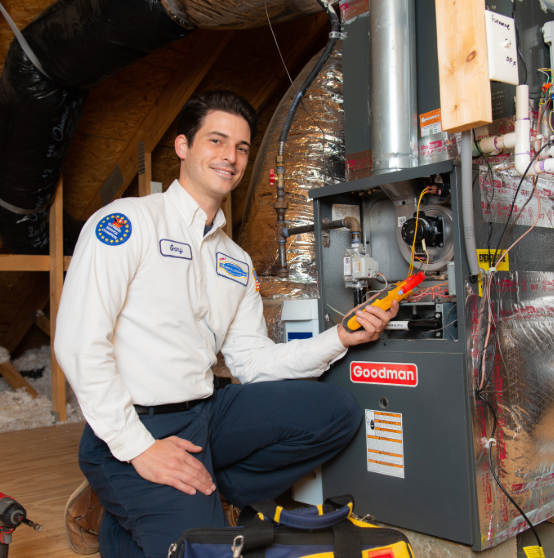 A technician in a uniform kneels beside an HVAC unit in an attic, smiling and holding a tool.