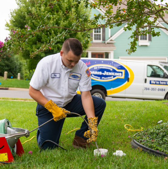 A technician in a uniform kneels on a lawn, using a plumbing tool. A service van is parked on the street in the background.