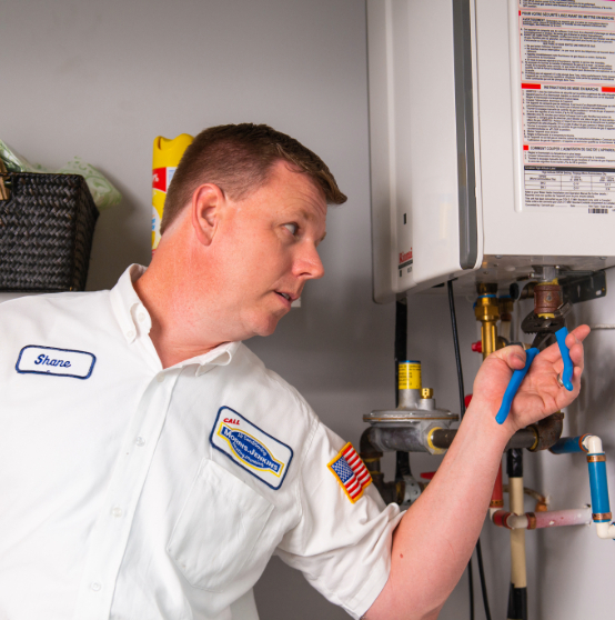 A plumber adjusts a valve on a wall-mounted water heater using a blue tool, wearing a white uniform with name and company patches.