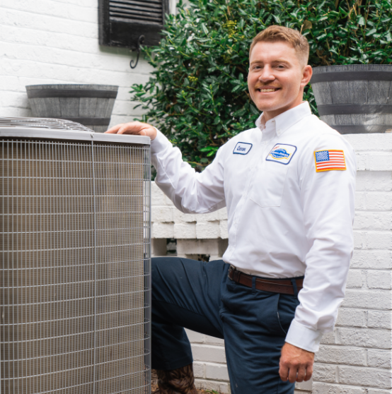 A technician in a white uniform with a name tag stands beside outdoor HVAC equipment, smiling, with one hand resting on the unit.
