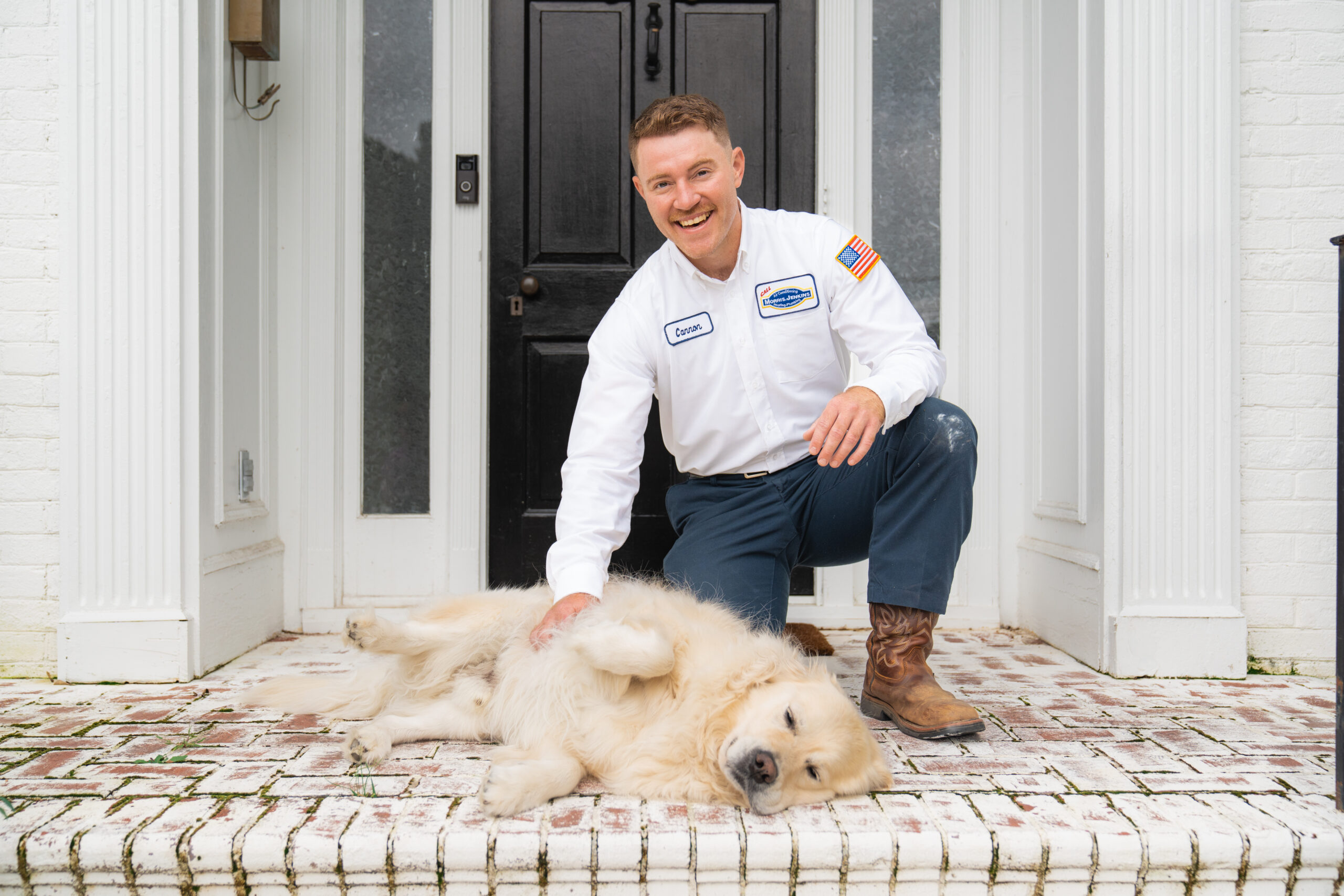 A person in a work uniform kneels on a porch, smiling and petting a large dog lying on its side.