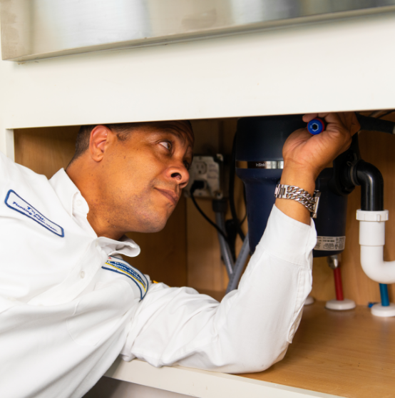 A man inspects plumbing under a sink, wearing a white uniform.