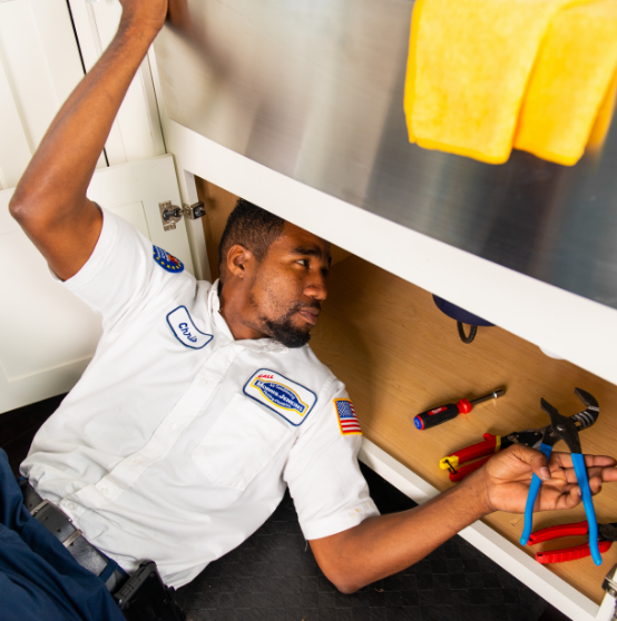 A technician in a uniform works under a kitchen sink, surrounded by tools including pliers, a screwdriver, and an adjustable wrench. A yellow cloth hangs above on the counter.