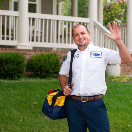 A uniformed technician stands on a lawn in front of a house, holding a tool bag and waving with a smile.