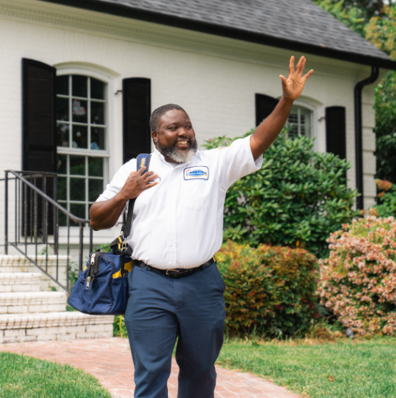 A smiling man in a work uniform waves and carries a tool bag while walking on a brick path in front of a house.