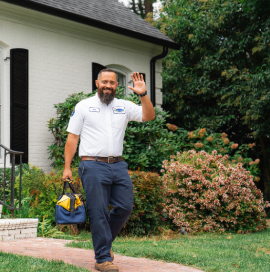 Man in a uniform holding a tool bag and waving while walking on a path in front of a house with green foliage.