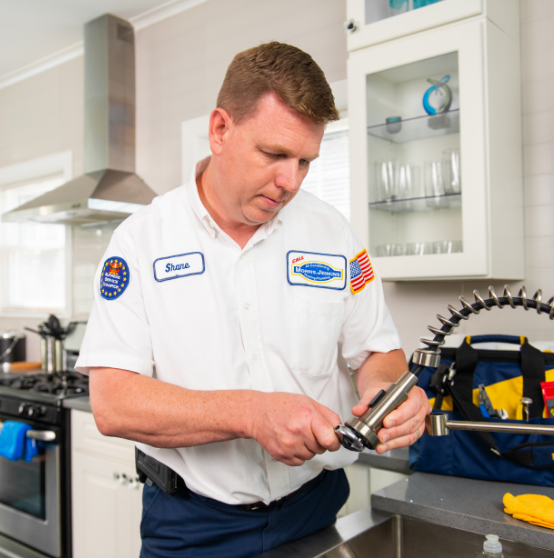 A plumber wearing a uniform labeled "Shane" repairs a kitchen faucet, with tools and a yellow cloth visible on the counter.