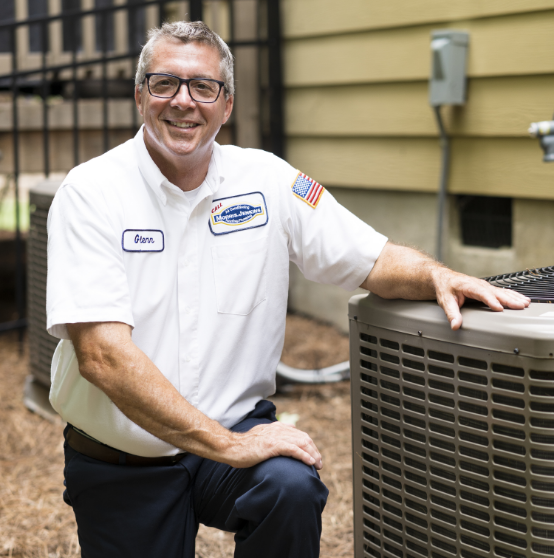 A maintenance technician in uniform kneels next to an outdoor air conditioning unit, smiling at the camera.